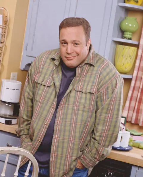 Kevin James in a plaid shirt, hands in his pockets, smirking in a sitcom kitchen