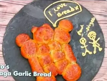 Mo's garlic bread Signature, golden rolls shaped into a muscled bear on a slate board, piped with "I love bread x" and stick figures of Paul and Nigella