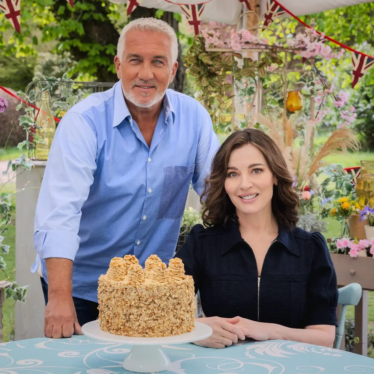 Paul Hollywood standing beside a seated Nigella Lawson behind a nut-covered layer cake, under Union Jack bunting in the Bake Off garden