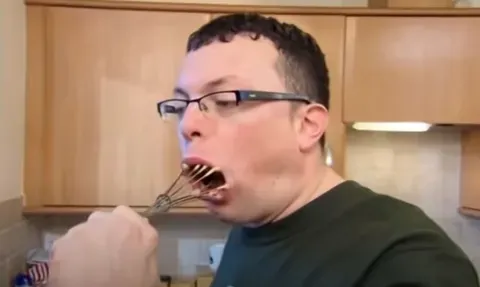 A man in glasses, mouth wide open around a whisk, licking it clean in a kitchen